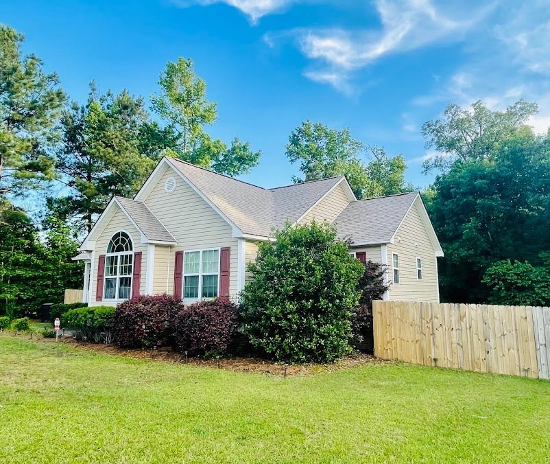 Single-story beige house with red shutters, front lawn, and a wooden fence under a blue sky.