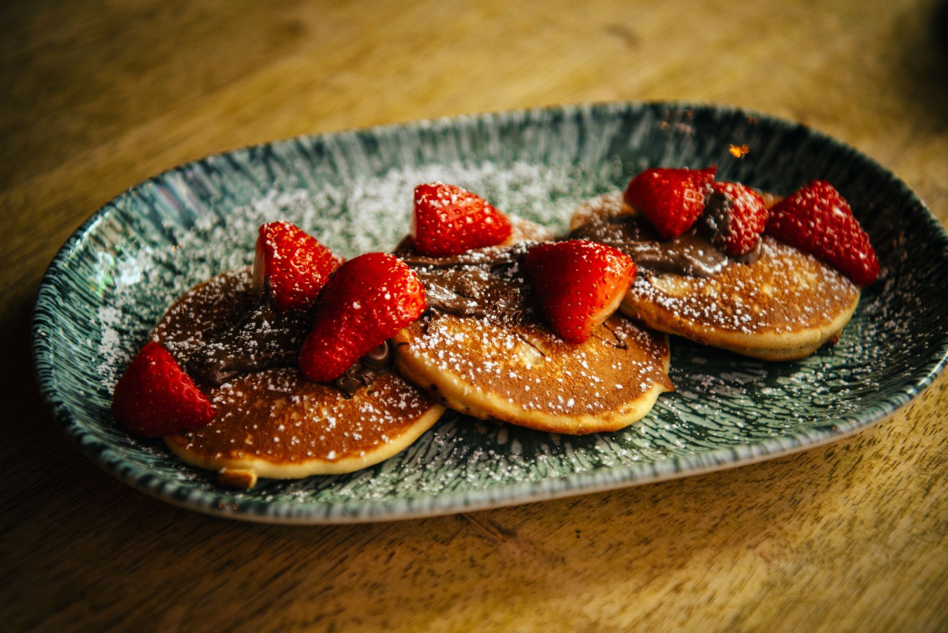Pancakes with strawberries and chocolate, dusted with powdered sugar, served on a textured plate.