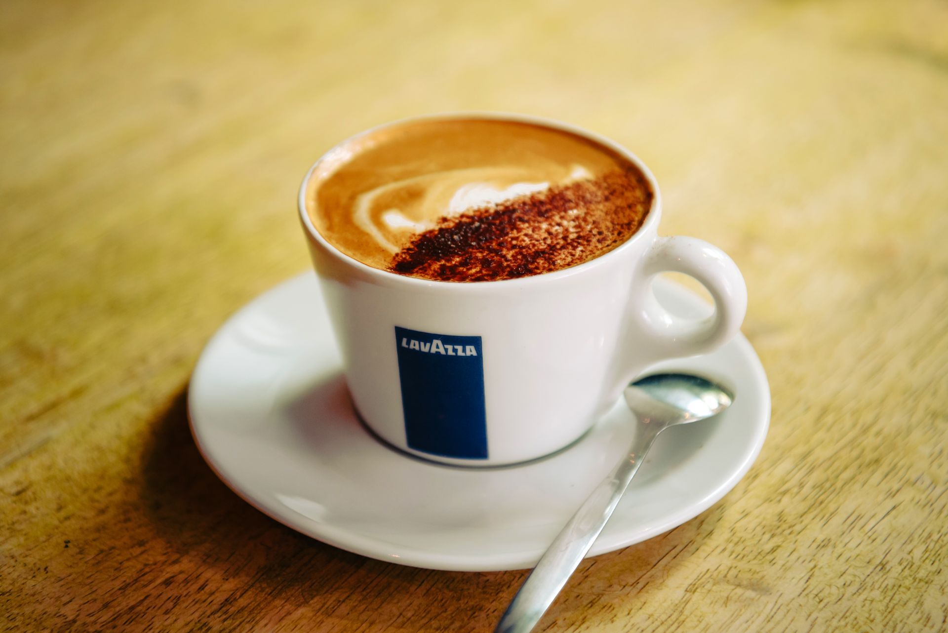 Cup of cappuccino on a saucer, with a spoon. Creamy coffee topped with cocoa powder, on a wooden table.