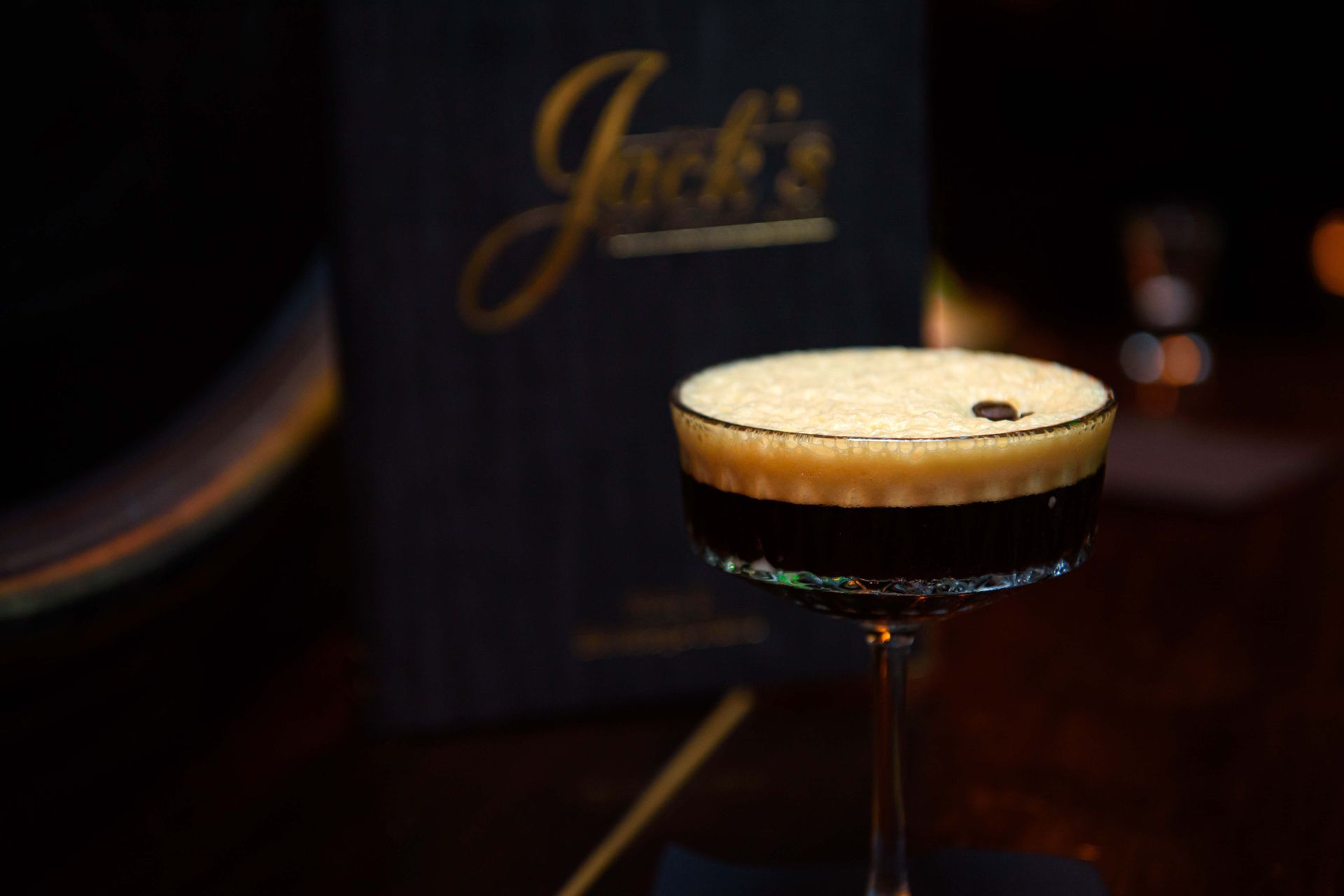 Espresso martini in a stemmed glass with foamy top, against a dark background, near a menu.