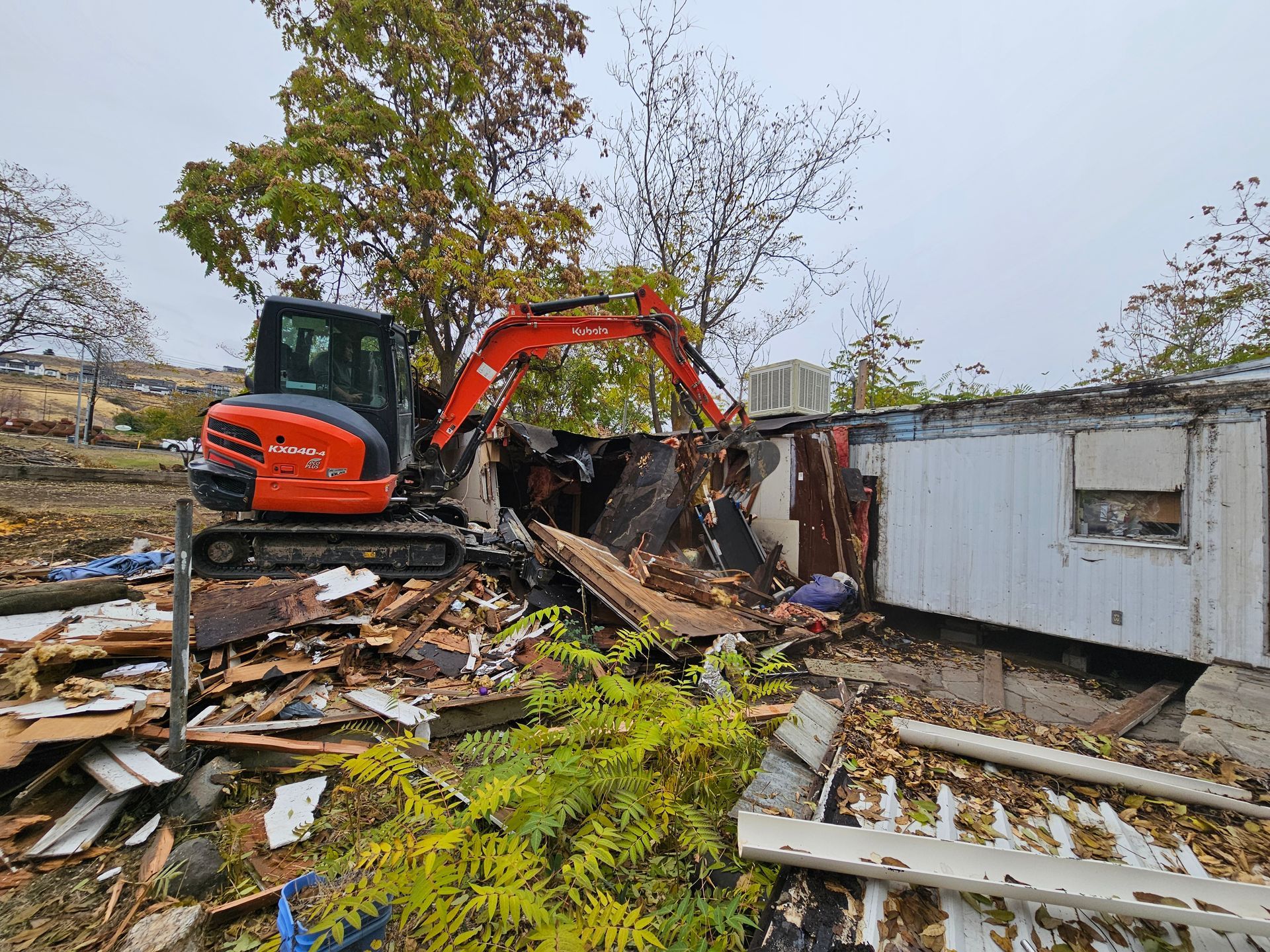 An excavator demolishes a mobile home, surrounded by debris. Orange machine, overcast sky.