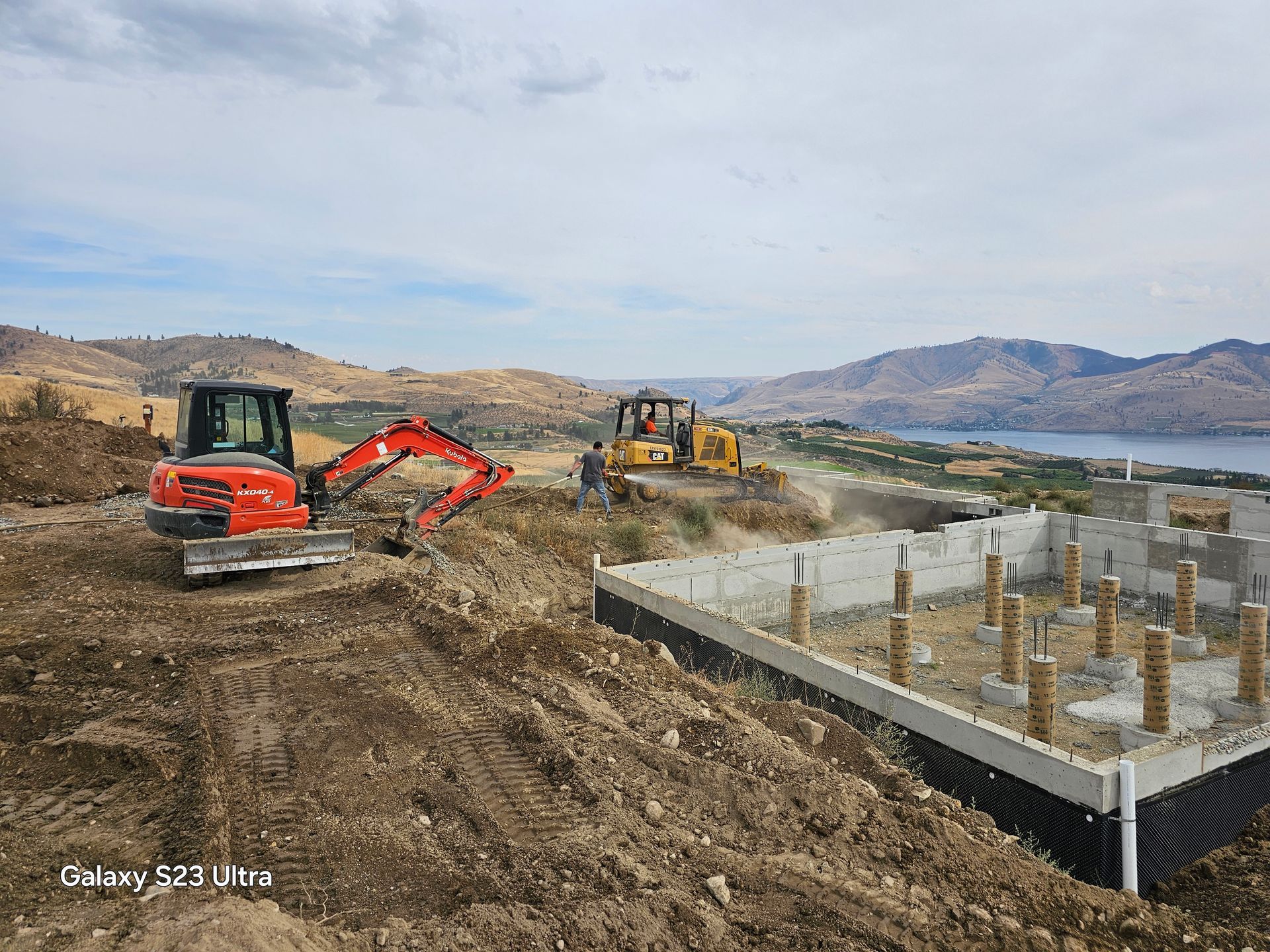 Construction site with excavator and bulldozer grading land near a concrete foundation, overlooking a lake and hills.
