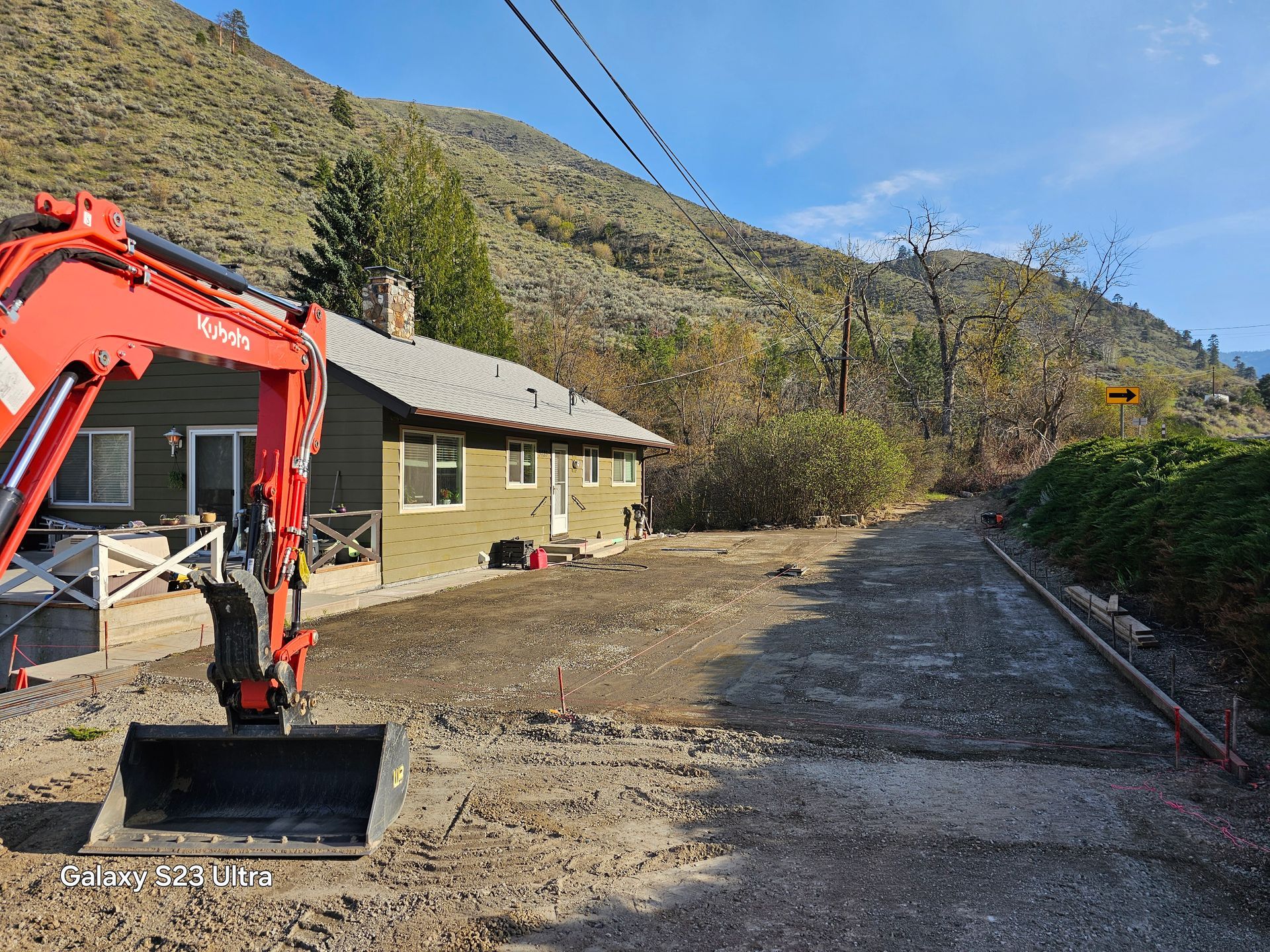 Excavator in front of a house, gravel driveway. Mountains in the background. Sunny day.