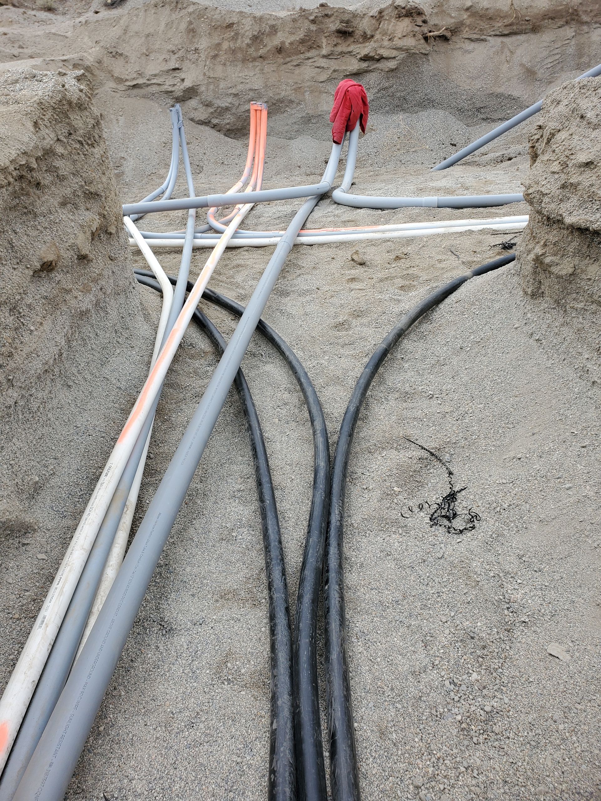 Pipes and wires in a trench, possibly for utilities. Some are gray, black, orange, and white; buried in gravel.