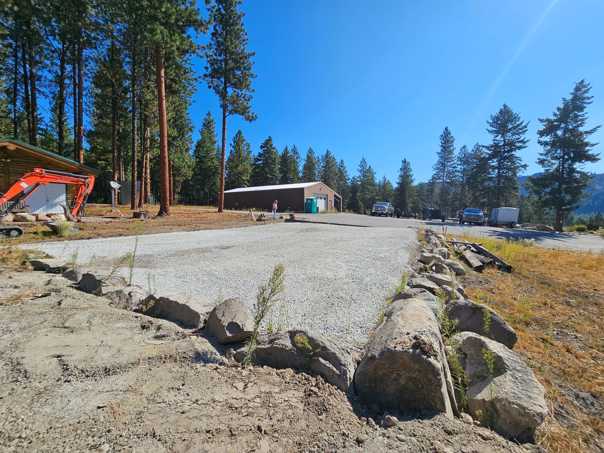 Gravel driveway bordered by large rocks, leading to buildings and vehicles, with trees and a blue sky in the background.