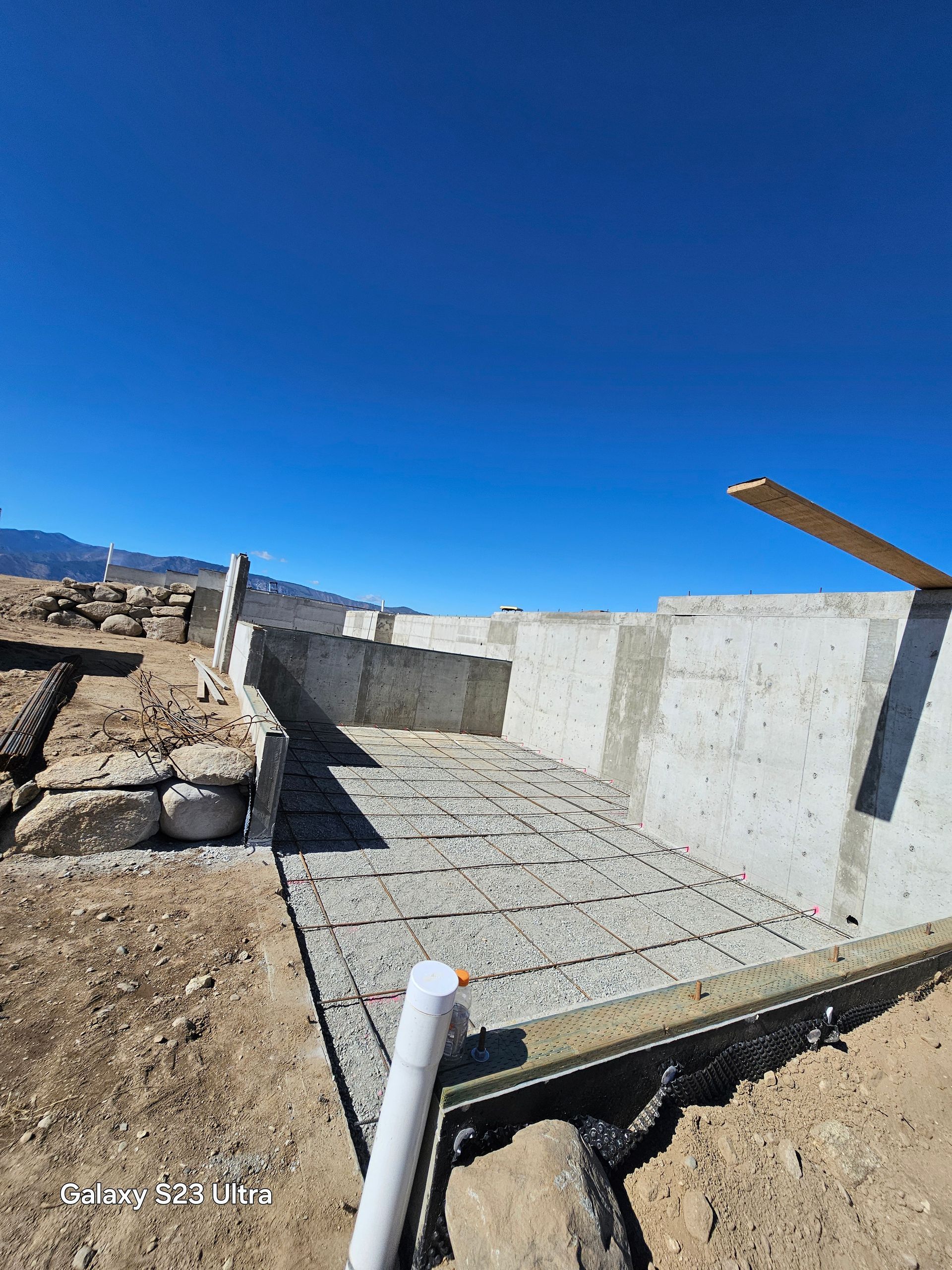 Concrete foundation construction site under a clear blue sky. Rebar grid and concrete walls visible.