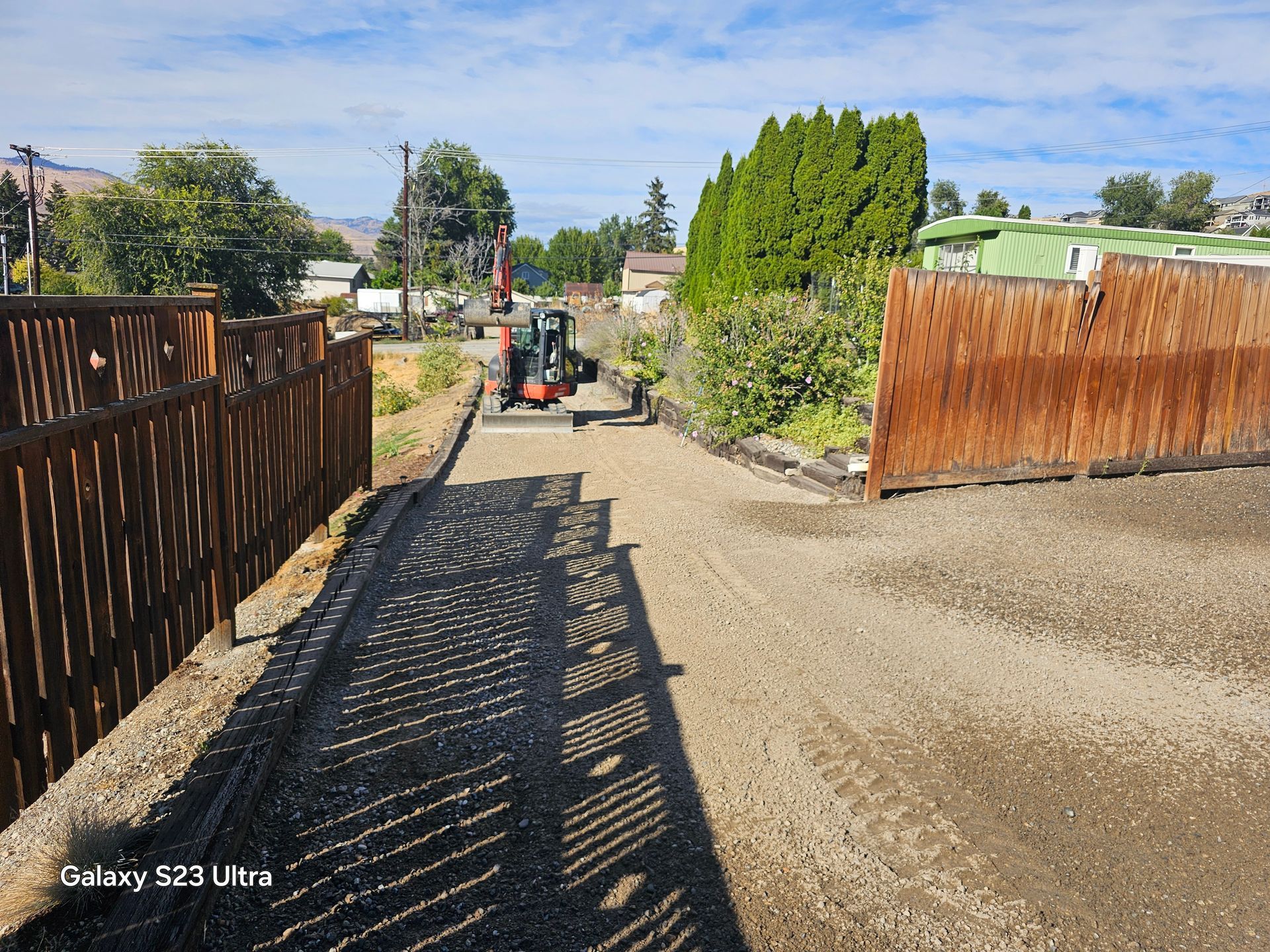 Construction site, small tractor paving gravel driveway. Wooden fences, blue sky, trees in the background.