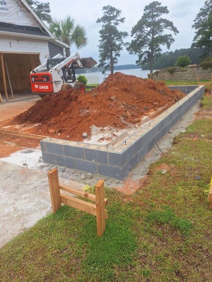 Construction site with excavator piling red dirt beside a house foundation and concrete block border Construction site with excavator piling red dirt beside a house foundation and concrete block border