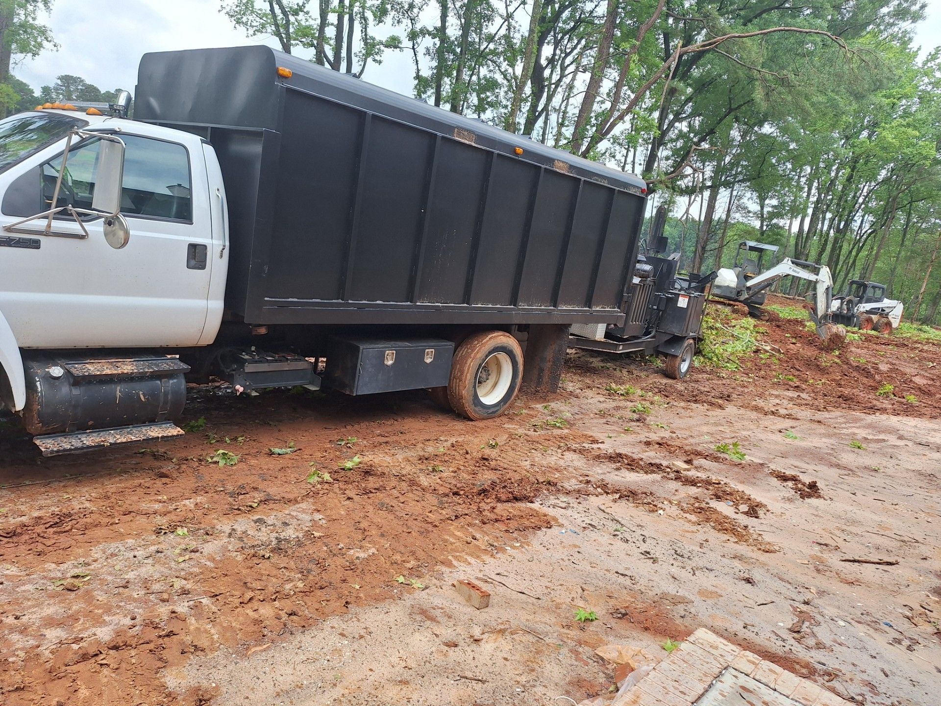 Black dump truck stuck in muddy dirt road, with a small excavator clearing brush nearby. Black dump truck stuck in muddy dirt road, with a small excavator clearing brush nearby.