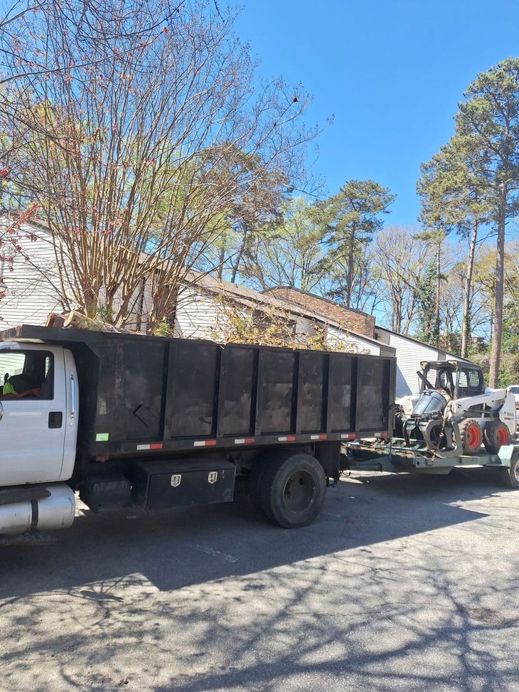 White truck hauling a black dump trailer and machinery on a sunny street beside leafless trees White truck hauling a black dump trailer and machinery on a sunny street beside leafless trees
