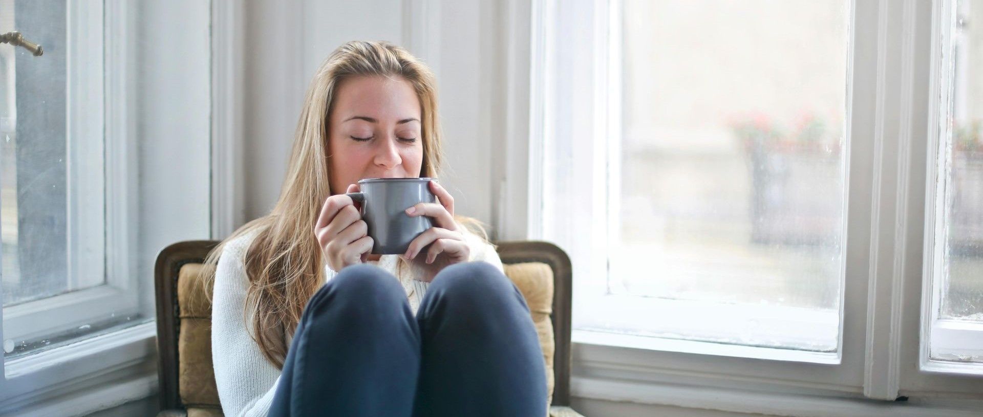 Woman sitting by a window drinking from a mug, wearing a blue sweater.