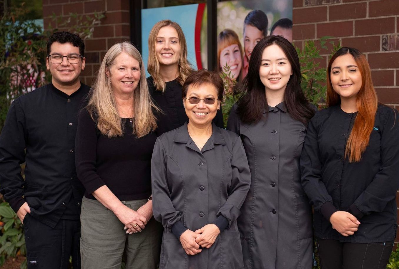 The Aurora Dental Care team standing outdoors in front of a brick wall, dressed in dark clothing and smiling.