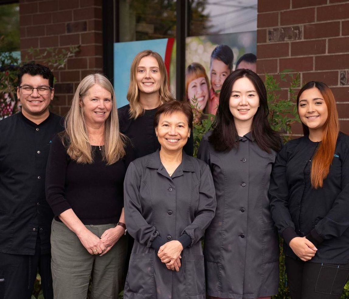 The Aurora Dental Care staff together outdoors in front of a brick building, wearing dark clothing and smiling.
