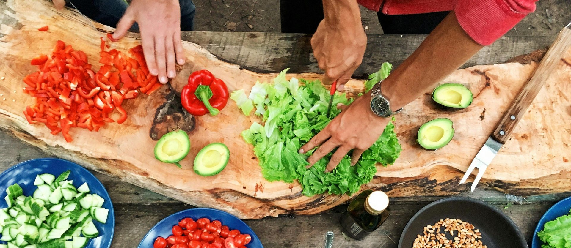 Hands chopping red peppers and green vegetables on a rustic table with avocados and bowls nearby