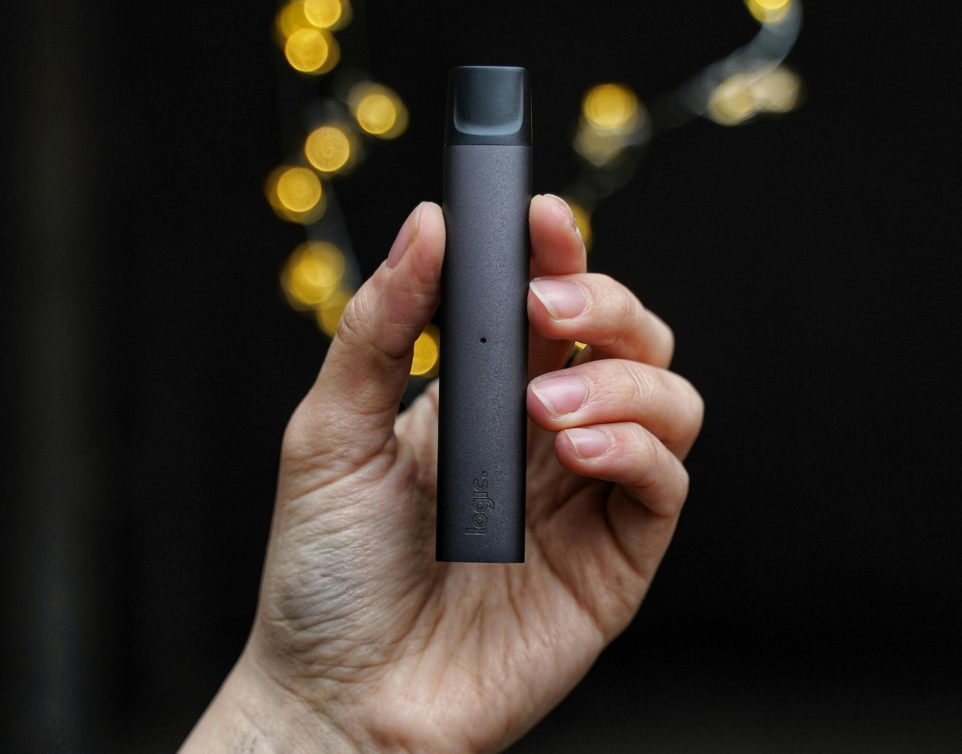 Hand holding a black vape pen against a dark background with blurred warm lights.