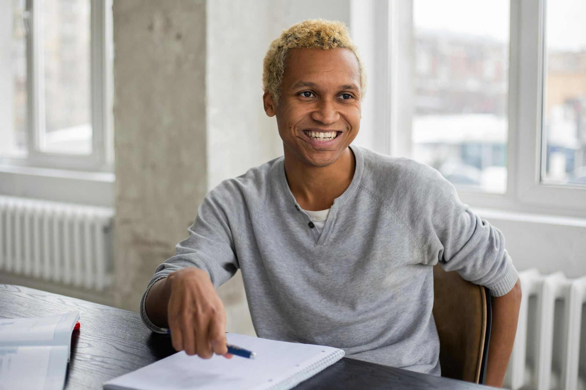 Smiling person in a gray shirt pointing at papers on a table by a bright window