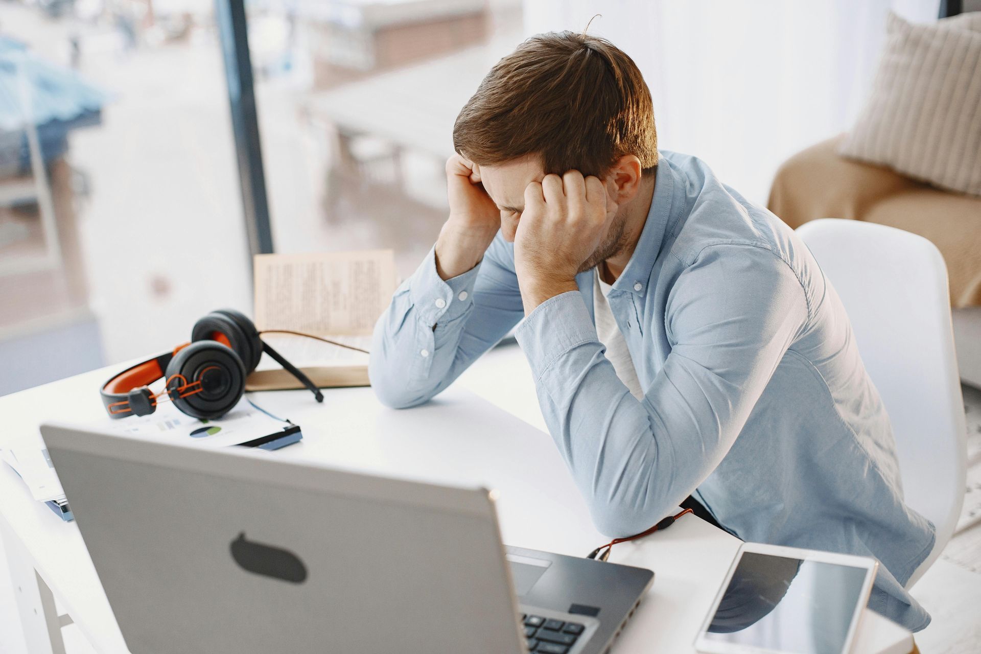 Stressed person at a desk, holding their head while looking at a laptop, with headphones nearby