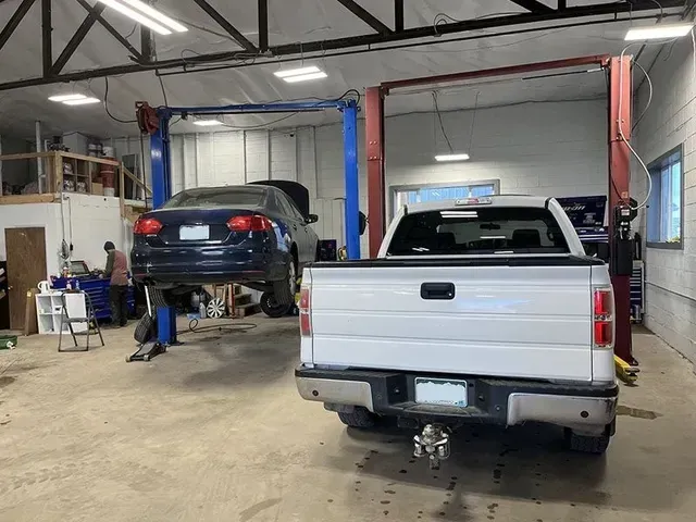 A white pickup truck and a dark sedan car in an auto repair shop, both raised on lifts. | Mohr's Autmotive
