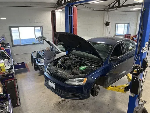A dark blue car raised on a lift in a repair shop with its hood open; a mechanic is working on it. | Mohr's Autmotive