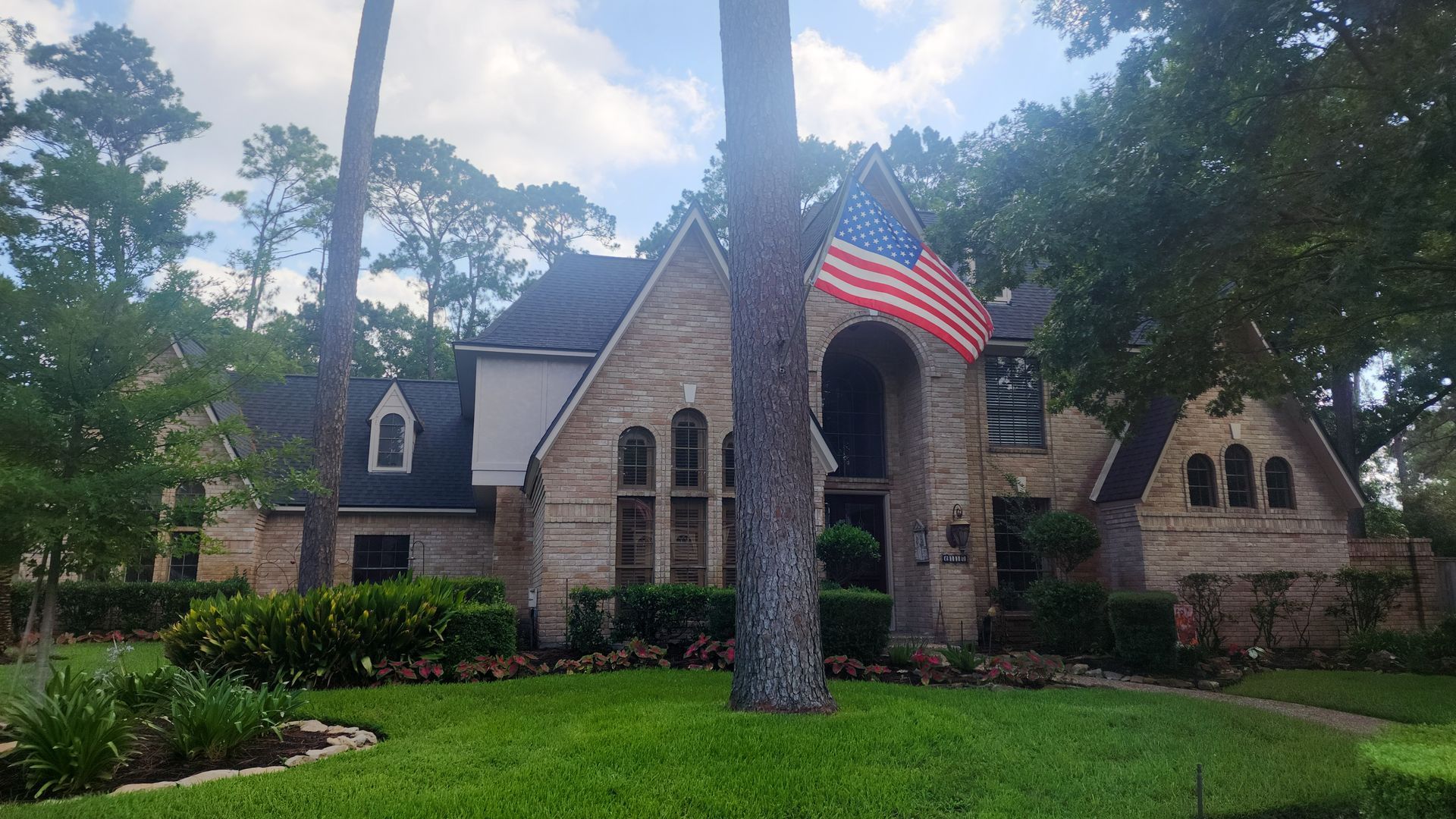 Stone house with arched entrance, large U.S. flag, and trees in a green yard under a partly cloudy sky