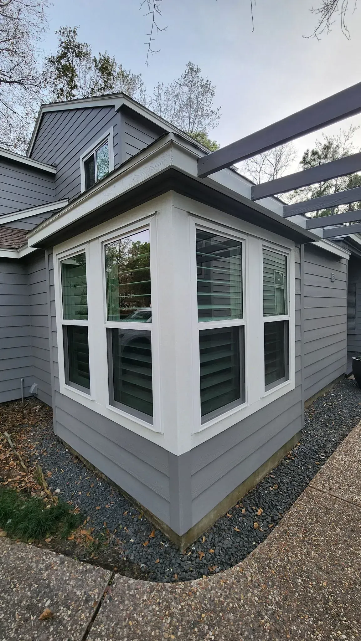 Gray house exterior with white-trimmed bay windows and a gravel path beside a patio structure