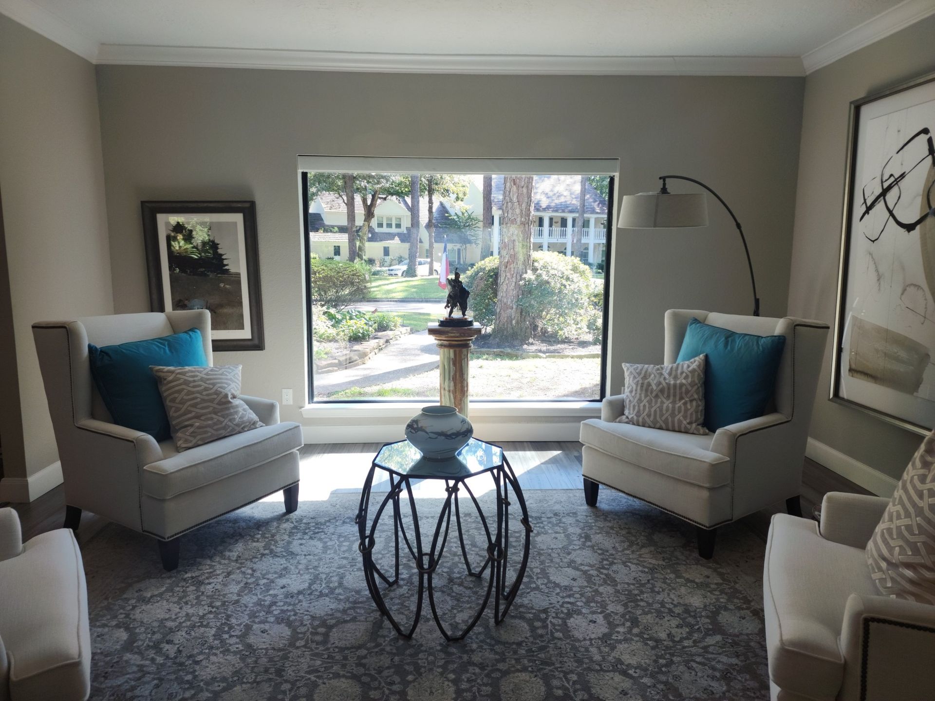 Elegant living room with two beige chairs, blue pillows, a round glass table, and a garden view window