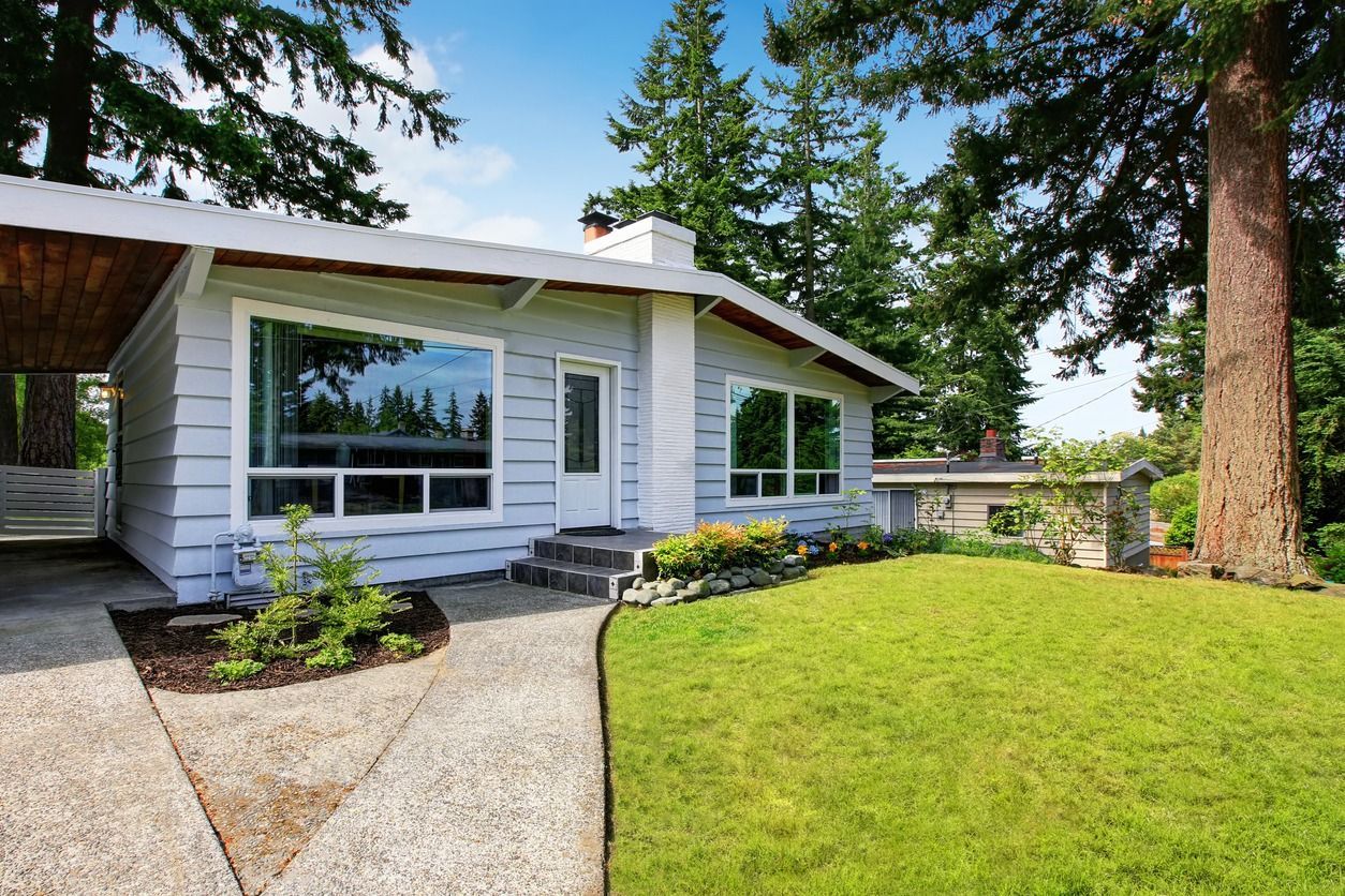 Single-story white house with a curved gravel path, green lawn, and tall trees in front.