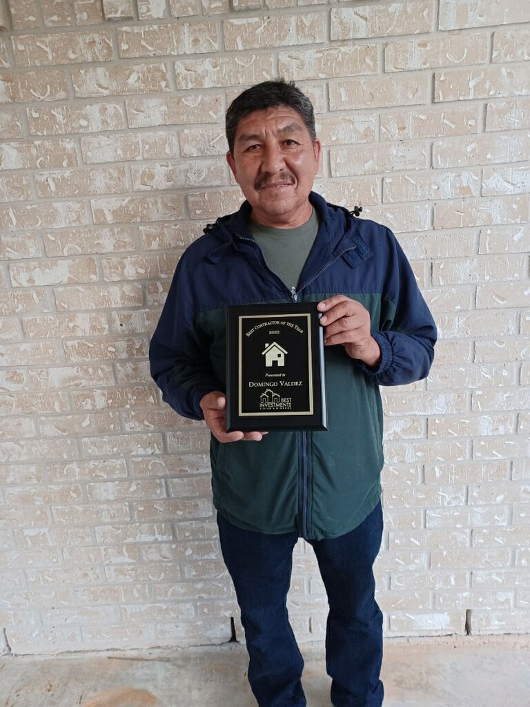Man standing against a brick wall, holding a framed plaque and smiling.