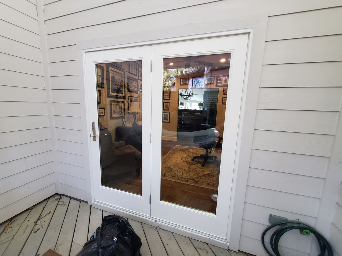 White French doors on a porch, with a black bag on the floor and a coiled hose by the wall.