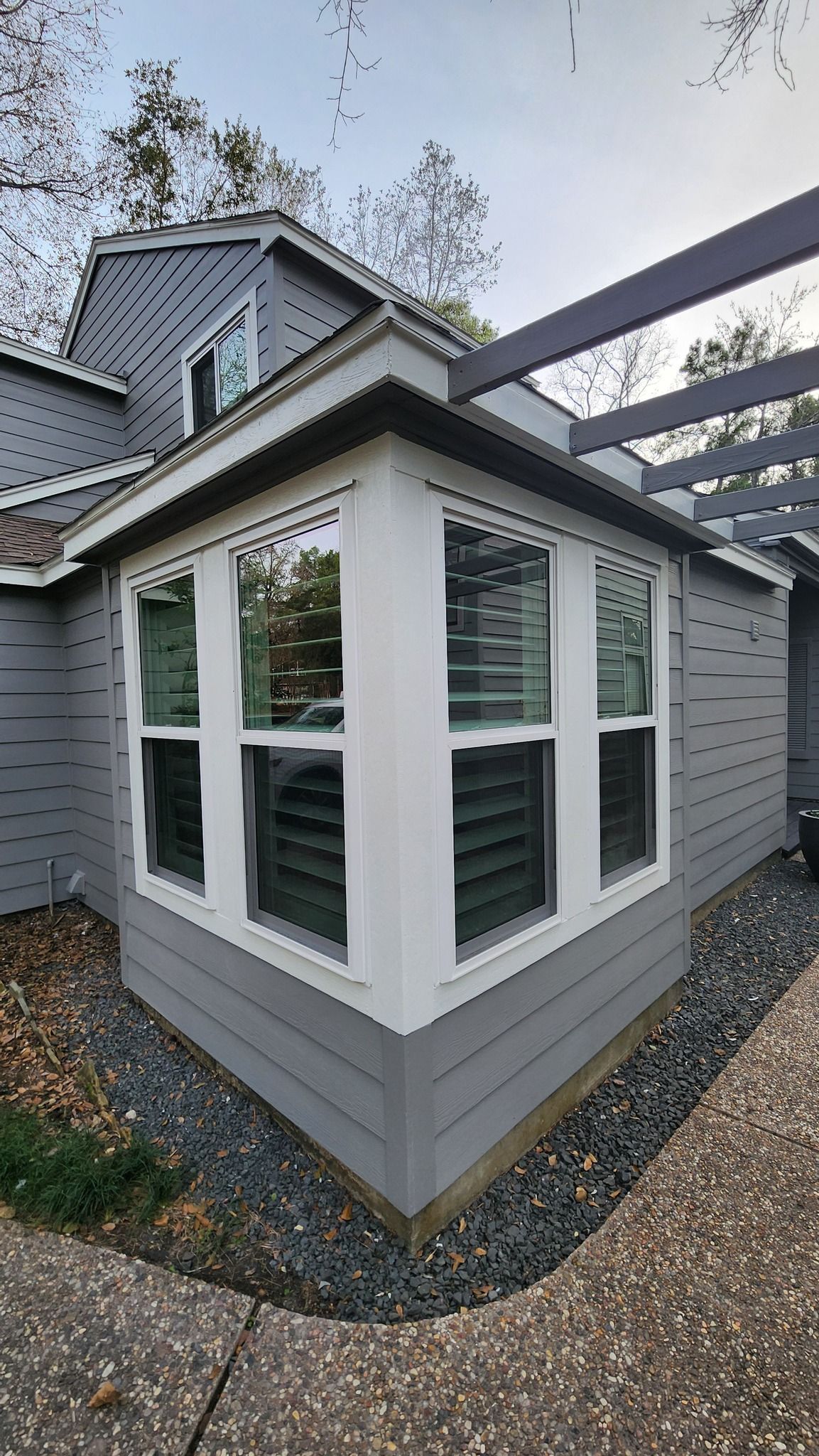 Gray house corner with white-trimmed bay windows and a sloped roof over a gravel path