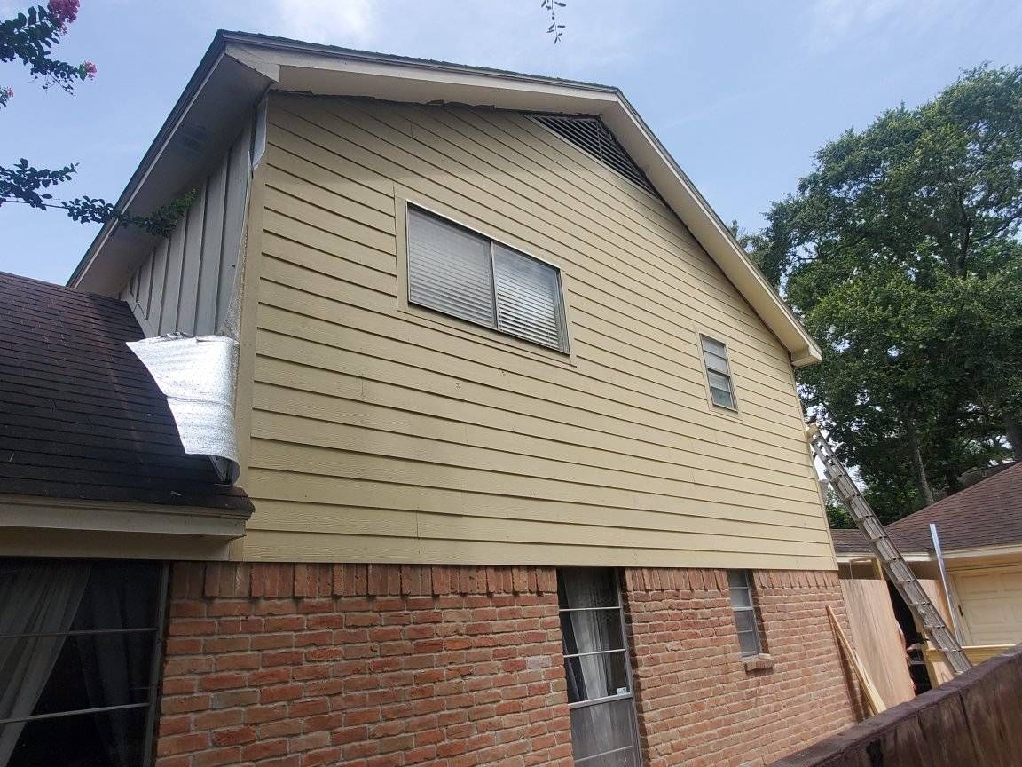 Two-story house exterior with tan siding, brick lower level, and a white-framed window under a blue sky
