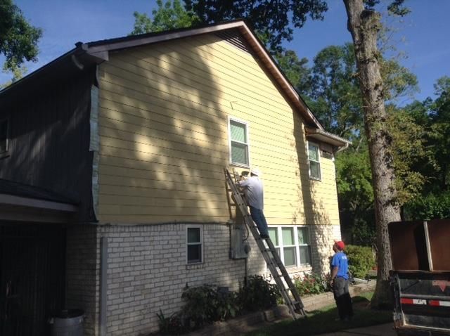 Two workers on a ladder painting the side of a yellow house beside a driveway and truck