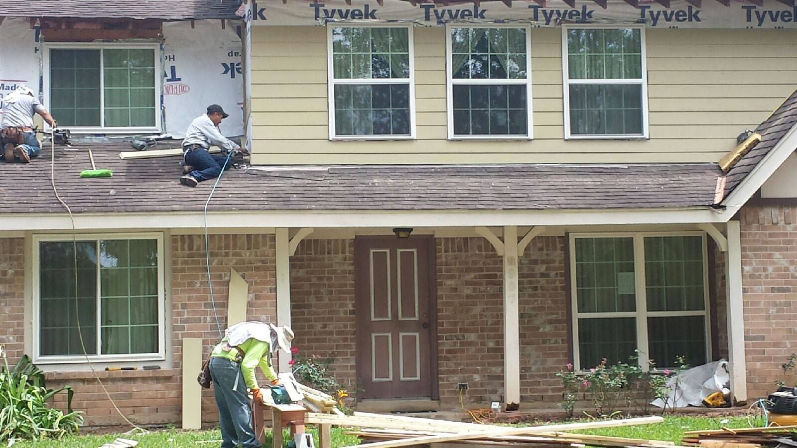 Workers repairing a two-story house roof and exterior siding with construction materials visible
