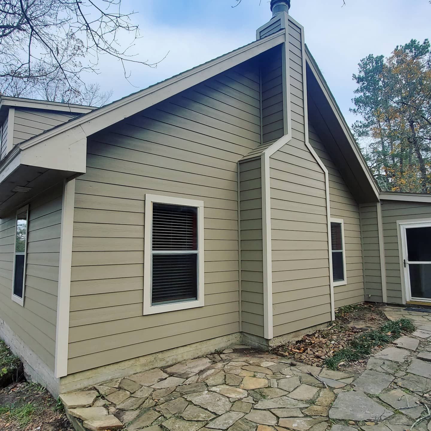 Side view of beige house with a stone walkway, gabled roof, and windows surrounded by trees