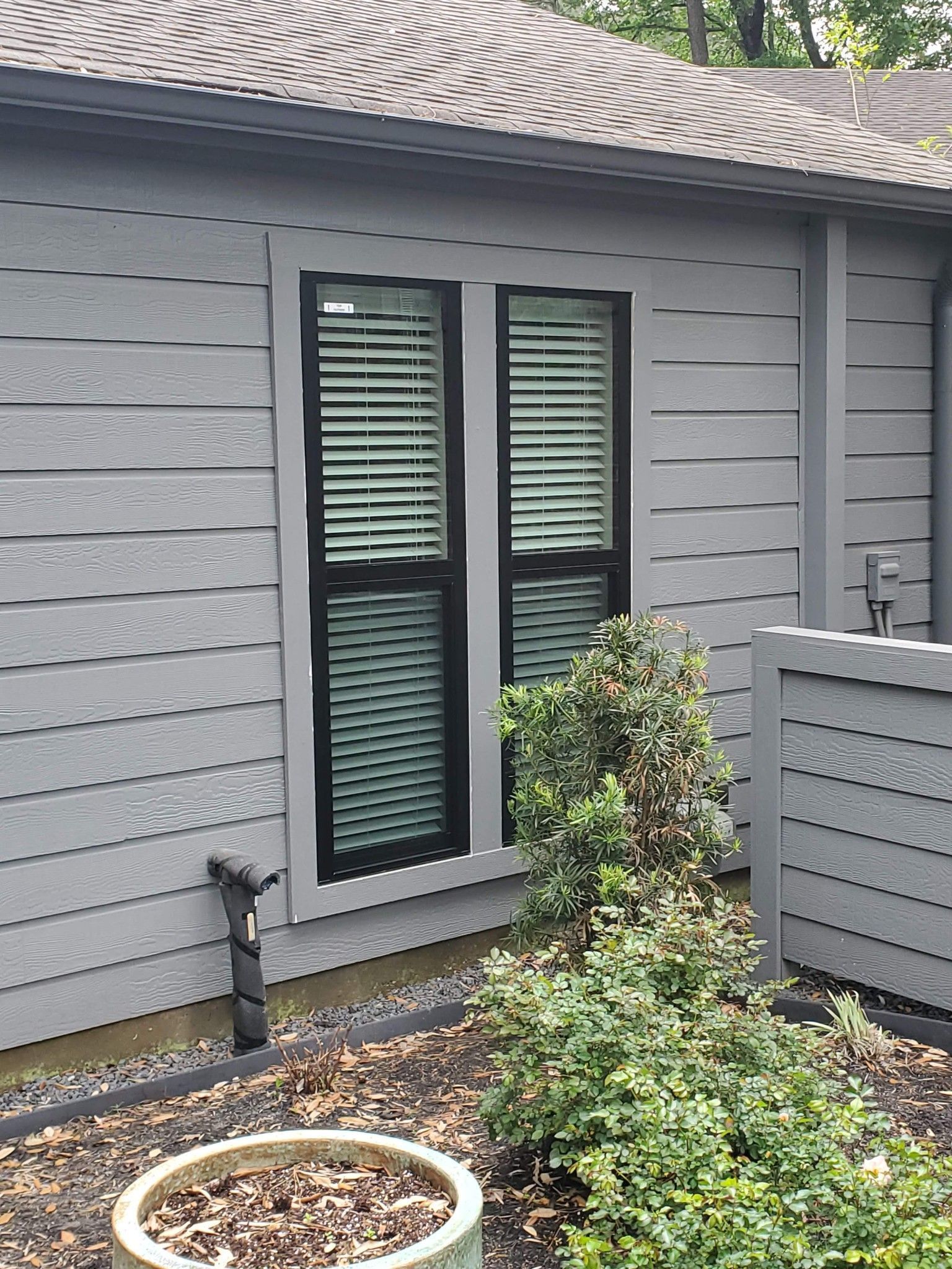 Grey house exterior with black-framed window, small bush, and gravel garden bed