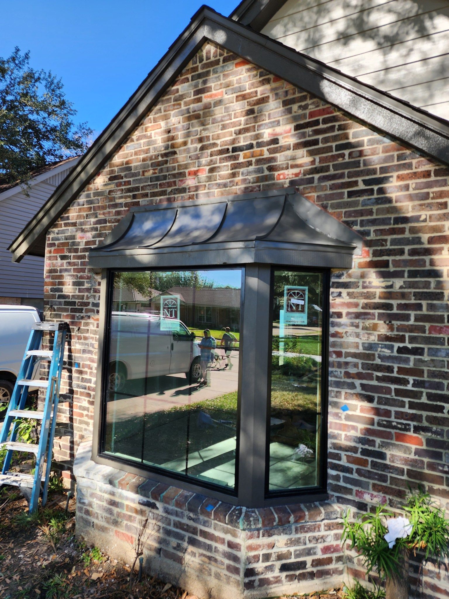 Brick house exterior with a black bay window and awning, next to a ladder in a sunny yard