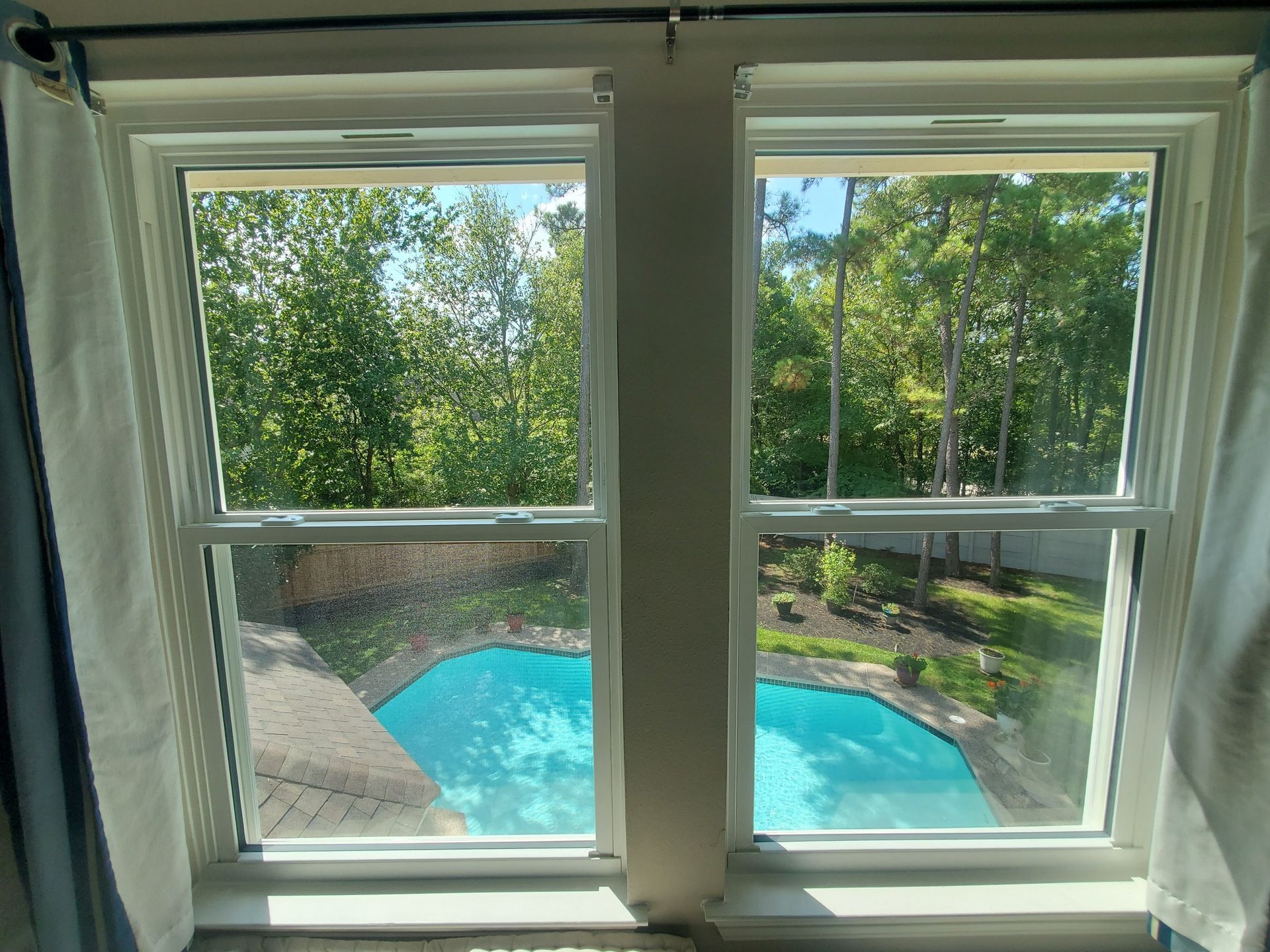 View through a window to a blue backyard pool surrounded by green trees and a stone patio
