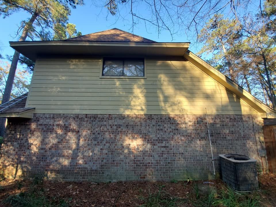 Side view of a house with brick lower walls and pale yellow siding in a wooded yard.