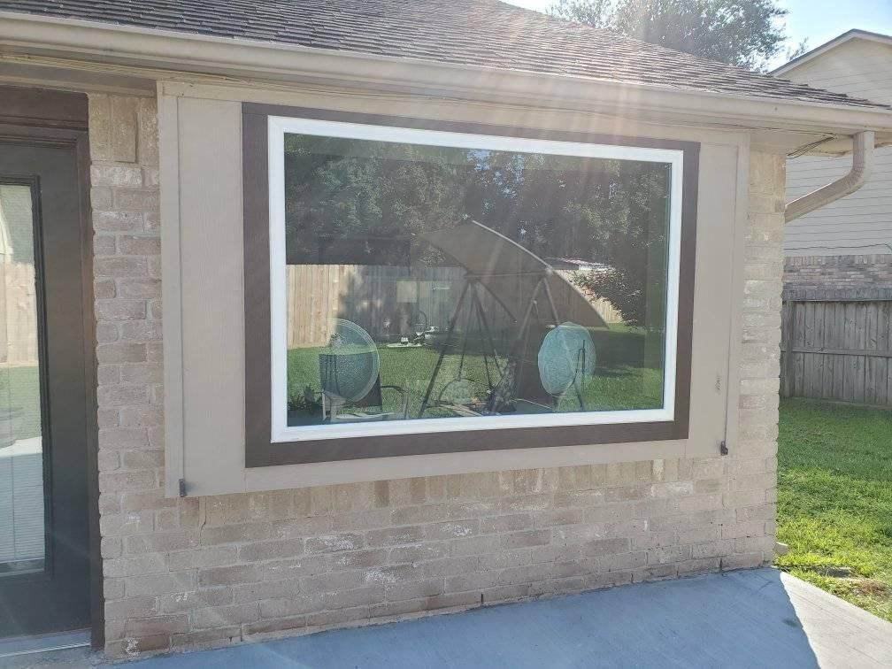 Large window with white trim on a brick house exterior, reflecting a backyard and patio chairs