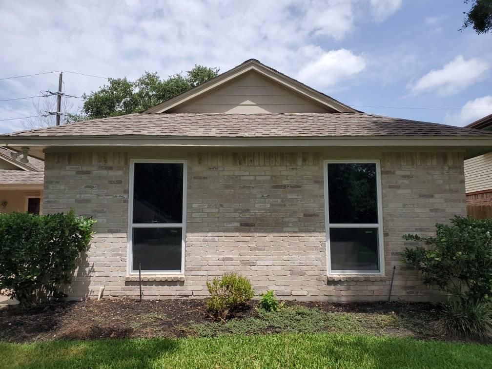 Front view of a beige brick house with two windows, a gabled roof, and shrubs in the yard