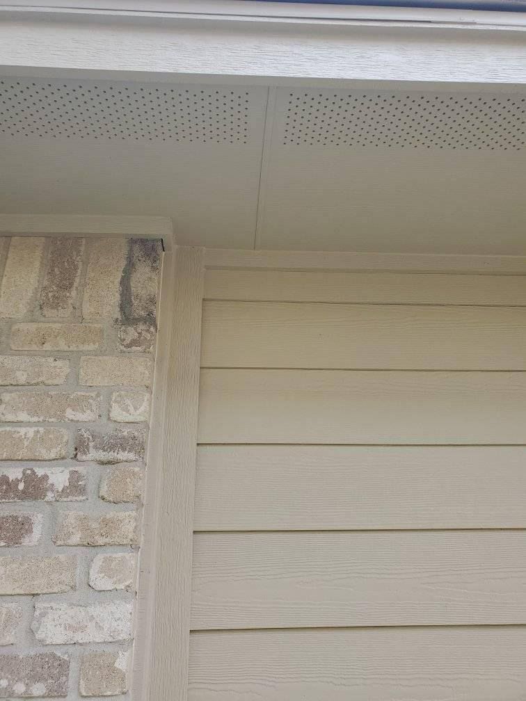 Close-up of beige house siding, brick wall, and soffit under the roofline.