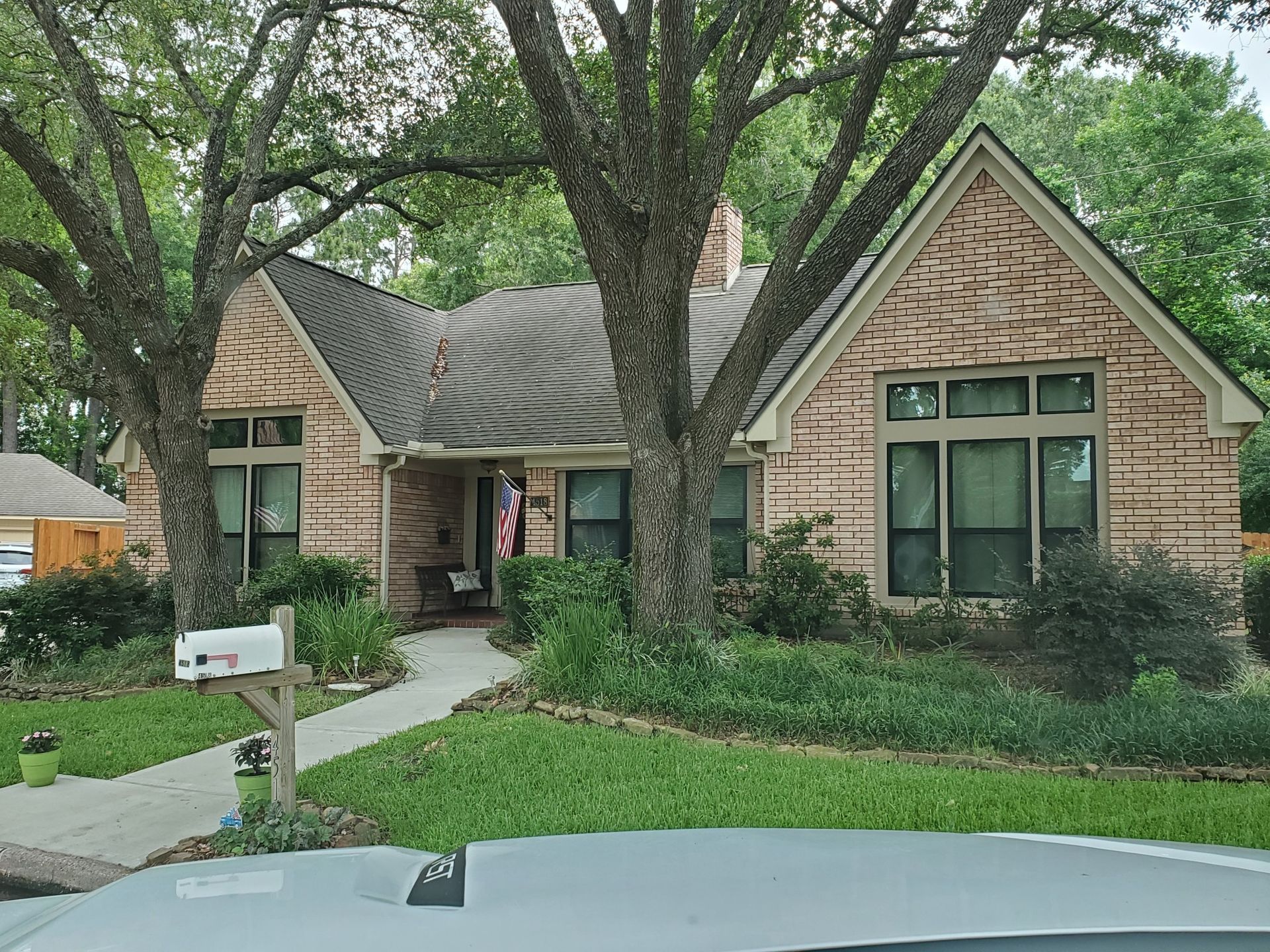 Suburban stone house with large trees, arched entry, and green lawn beside a driveway and mailbox.