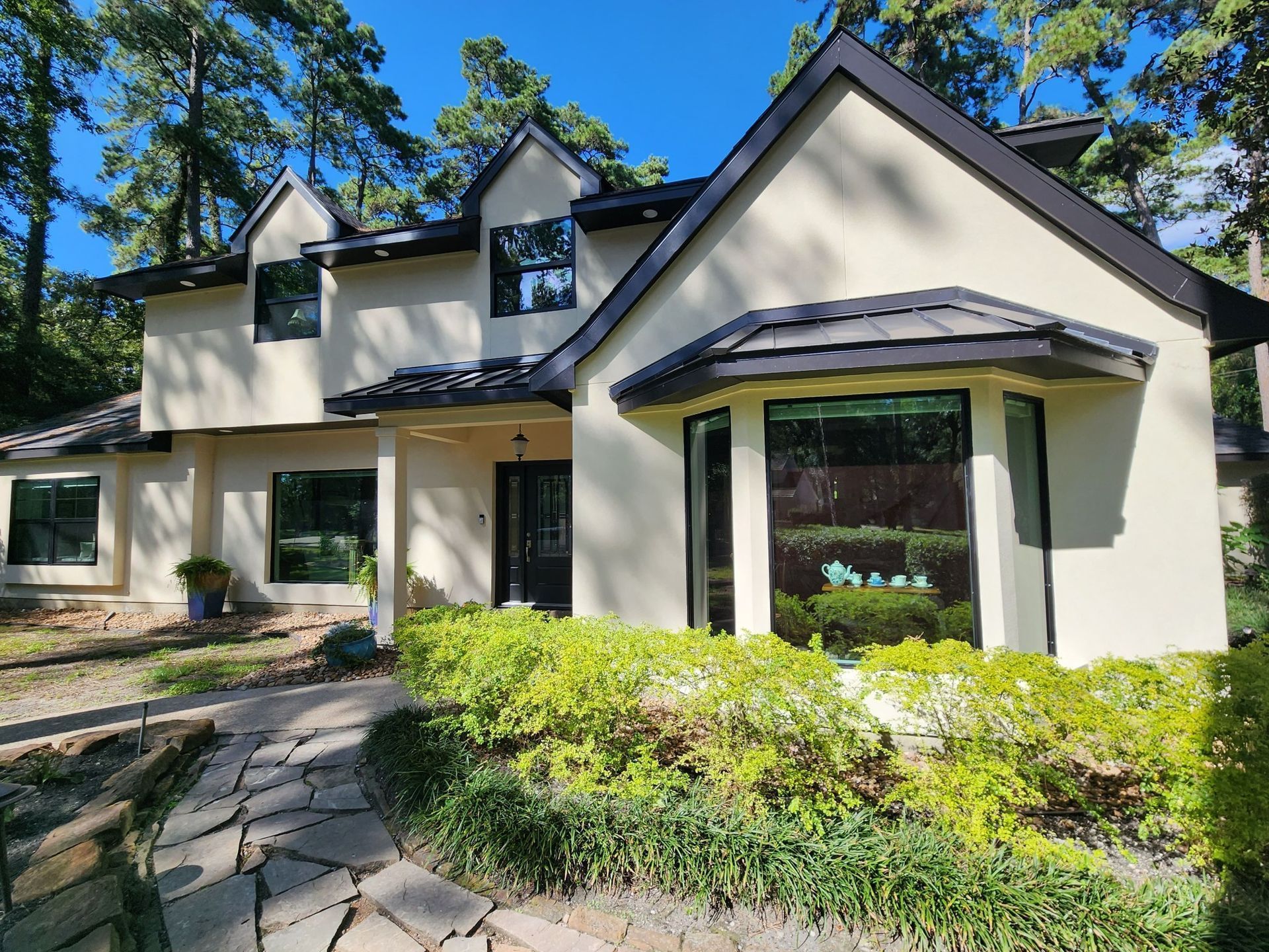 Cream-colored house with black trim, large windows, and a curved stone walkway surrounded by greenery