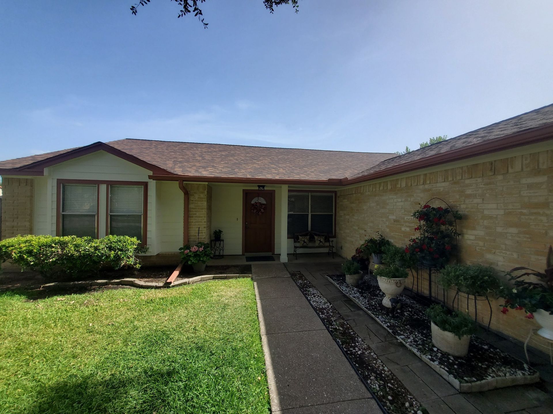 Single-story beige house with a red roof, front walkway, lawn, and small shrubs under a blue sky.
