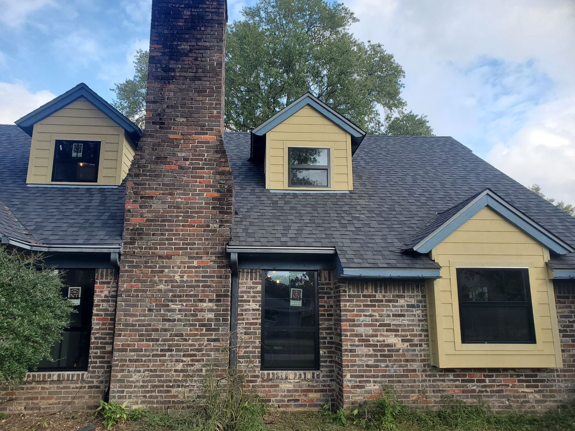 Brick house exterior with a large central chimney, dark shingle roof, and yellow-trimmed dormer windows