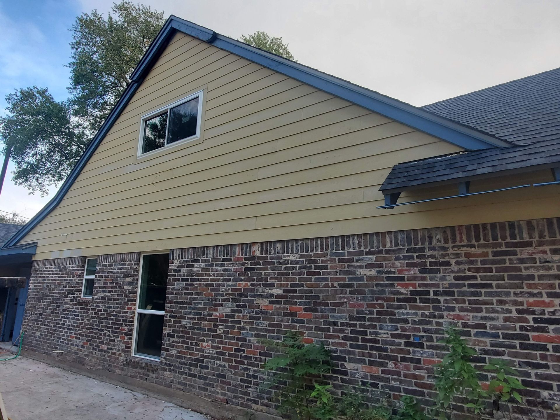Side view of a brick house with tan siding, a small window, and dark blue roof trim