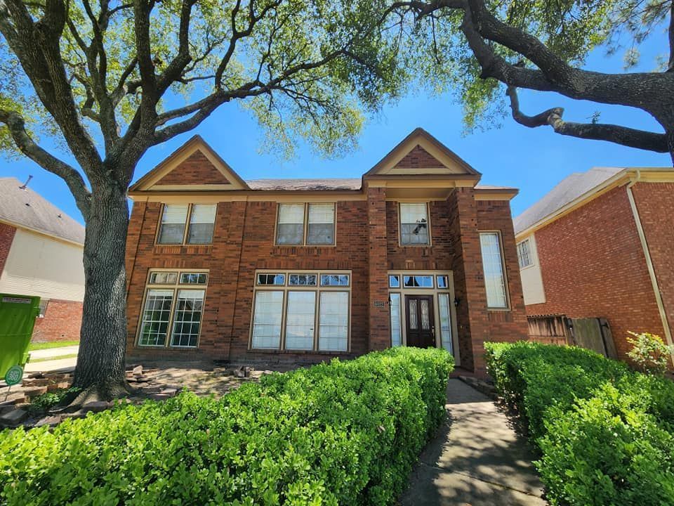Brick two-story townhouse with white-trimmed windows, a front walkway, hedges, and large trees overhead