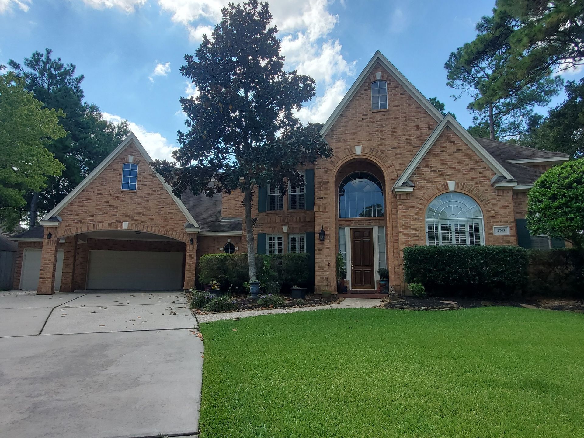 Brick suburban house with a two-car garage, arched entryway, and green lawn under a partly cloudy sky