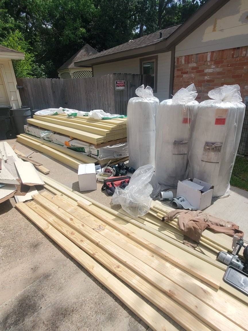Stacked lumber and packaged insulation materials on a driveway beside a house under construction.