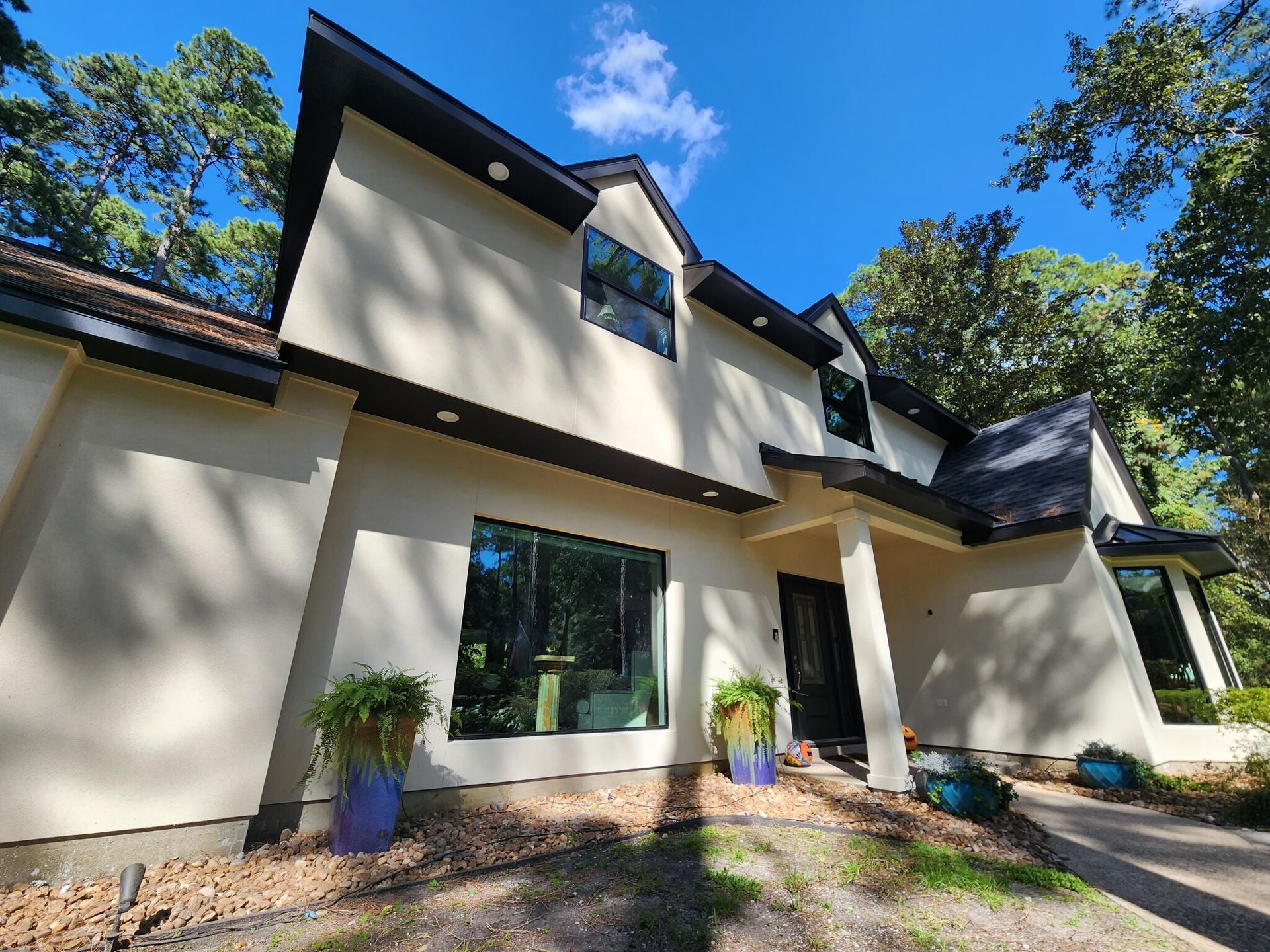 Modern cream-and-black house exterior with large windows, potted plants, and trees under a blue sky